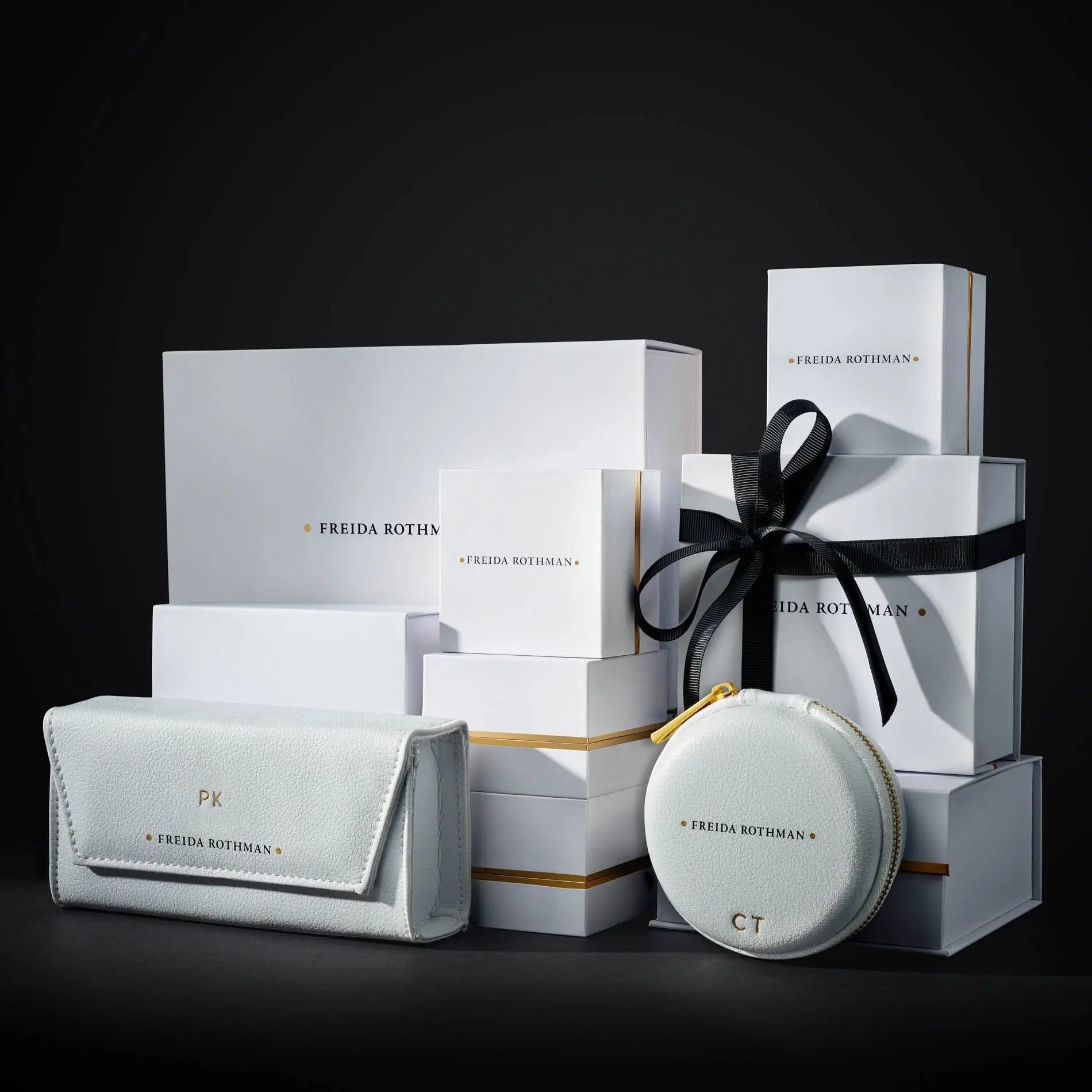 A stack of white gift boxes with gold-trimmed edges and a subtle rhodium matte finish, elegantly labeled "FREIDA ROTHMAN," against a dark background. In front, there&