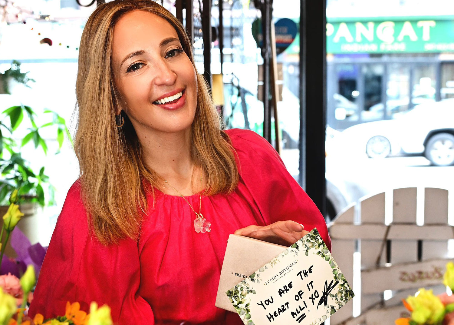 Freida in a red shirt holding a card with text, surrounded by flowers and blurred outdoor background